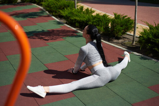 A sporty woman in a tracksuit sitting on the sports playground and performing a transverse twine. Back view. Sports, stretching.