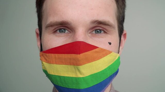 Close-up portrait of a young man with a black heart on his cheek taking off a rainbow face mask. A gay man in a rainbow LGBT pride mask.