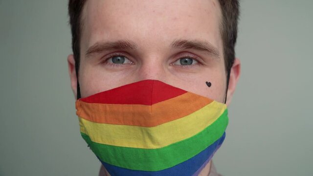 Close-up portrait of a young man with a black heart on his cheek wearing a rainbow face mask. A gay man in a rainbow LGBT pride mask.
