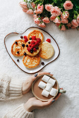 Woman holding a cup with cocoa and marshmallows, flowers and a plate with cheesecakes and berries. Close up. Breakfast concept
