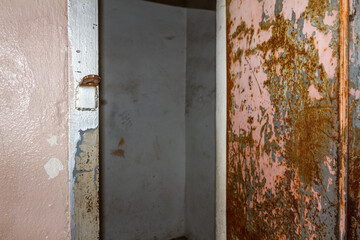 Open door within a rusted metal container type wall and paint chipped and faded entrance. Grey back wall and pealing pink and white paint. 