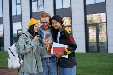 Emotional friends using mobile phone, watching videos online. Multiracial smiling students communication, talking, standing in university campus. Group of diverse people concept 