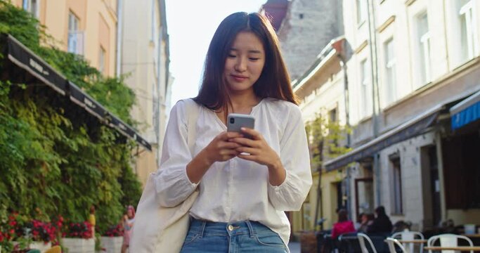 Cheerful female student going home after hard studying day and using smartphone. Young Asian woman walking down city street, texting message and smiling. Communication, technology concept.