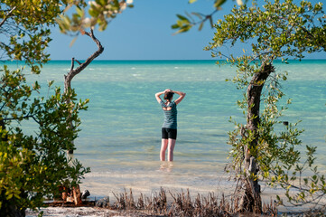 Young woman on the sandy beach by the sea in Holbox Island wearing casual summer clothing with her hands on her head. in the background the trees and the Caribbean Sea