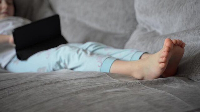 A little girl lies at home on the couch and looks into the black tablet. The child plays and watches the video. The girl has bare feet. The child is wearing pajamas. Close up. 4K