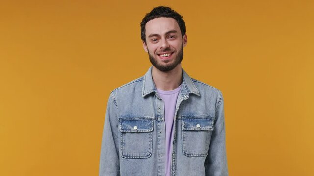 Handsome unshaven bearded young man 20s in denim jacket purple t-shirt looking camera asides charming smile isolated on yellow color wall background studio portrait People emotions lifestyle concept