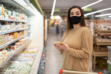 Close-up portrait, a young lady in a supermarket stands near a shop window, a protective mask is put on her face. Pandemic shopping concept.