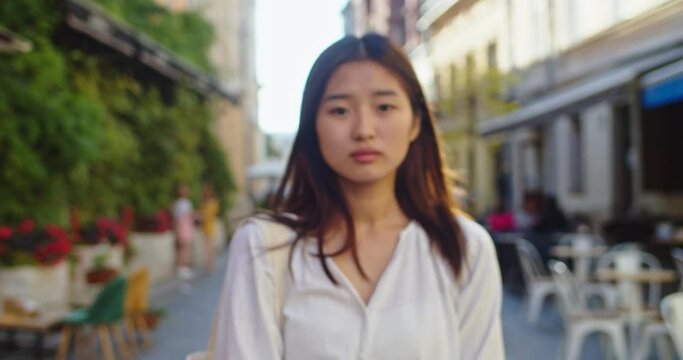 Portrait of beautiful Asian woman looking at camera and smiling. Young lady with wing in her hair wearing casual outfit, standing in urban city street outdoors. Walking, summer concept.