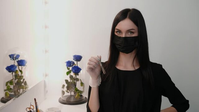 Beautiful makeup artist in a protective mask on her face in a beauty salon holds tweezers. Cosmetology service concept