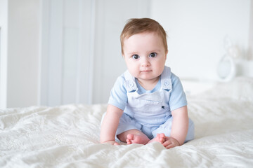 Fototapeta premium baby boy 6 months old in blu clothes smiling and sitting on white bed at home.