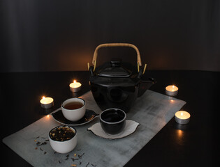 Beautiful black and white tea ceremony set: teapot and small cups and saucers next to a cup filled with dried tea leaves with pieces of citrus on the background of a black table with burning candles