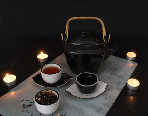 Beautiful black and white tea ceremony set: teapot and small cups and saucers next to a cup filled with dried tea leaves with pieces of citrus on the background of a black table with burning candles