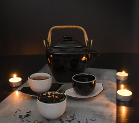 Beautiful black and white tea ceremony set: teapot and small cups and saucers next to a cup filled with dried tea leaves with pieces of citrus on the background of a black table with burning candles