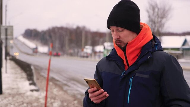 A man on the side of the dorga dials a number on his smartphone. A male in a warm jacket with an orange collar stands on the highway. In the background, cars pass in out of focus.