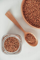 Buckwheat kernels in a glass jar, wooden bowl and spoon on white background. Top view. Copy, empty space for text