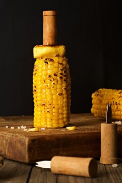 Melting butter on a corn on the cob standing on a wooden board against a black background and corn holders in the foreground.  