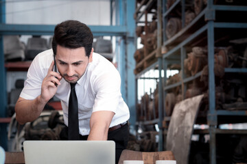 Portrait of smiling businessman seller working with laptop computer and talking on mobile phone at auto spare parts store shop warehouse with many second hand engine parts as blurred background.