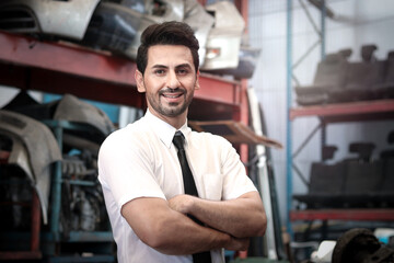 Portrait of smiling businessman seller standing with arms crossed at auto spare parts store shop warehouse with many second hand engine parts as blurred background.