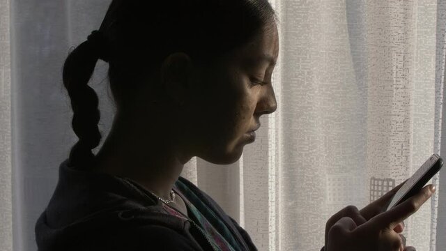 Young Ethnic Female Teenager Texting On Mobile Phone Near Window Curtains. Locked Off