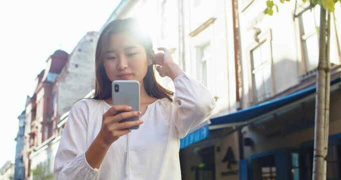 Charming female student using smartphone and standing in urban city street. Young Asian woman texting messages, browsing and smiling. Social media, technology, communication concept.