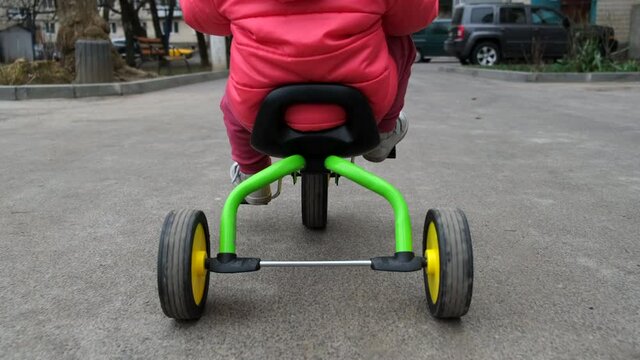 Following for little girl who rides a tricycle down the street. Back view.