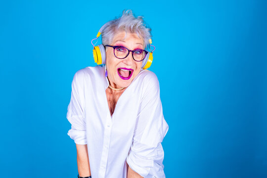 Old senior woman enthusiastic dancing listening music ecstatic isolated on blue background enjoying streaming playlist wearing bluetooth wireless headphones