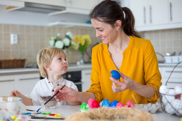 Beautiful blond child, toddler boy, painting easter eggs with mother at home, making easter wreath