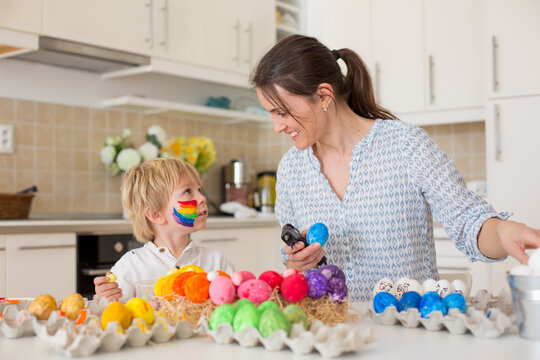 Beautiful blond child, toddler boy, painting easter eggs with mother at home, making easter wreath