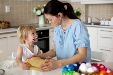 Little blond toddler child with painted face as rabbit and young mother, preparing dough for easter brioce buns, sweet easter bread