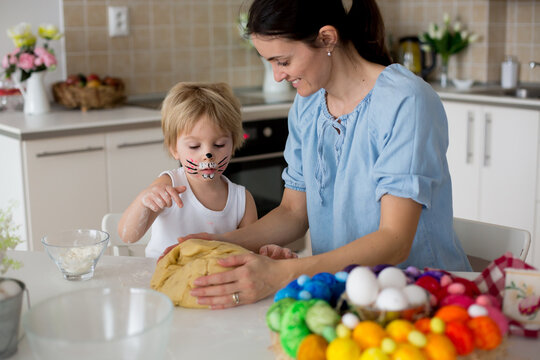 Little blond toddler child with painted face as rabbit and young mother, preparing dough for easter brioce buns, sweet easter bread