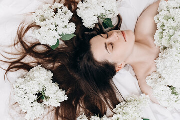 Naklejka premium Top view beauty portrait of sexy brunette girl lying on comfy white bed near small beautiful flowers. Slender young woman, perfect clean skin and long hair. Natural cosmetics for skin care.