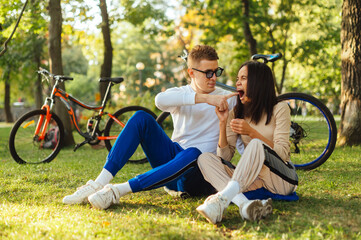 Obraz premium Funny young couple is laughing and joking, when sitting on a grass in the park. Two people having fun, a man is pointing a finger to a woman's mouth, a female is trying to bite a finger.