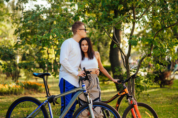 Obraz premium Attractive couple of young adults is in the city park with their bikes, man embracing a woman. Couple of cyclists is outside feeling carefree and romantic.