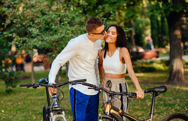 Obraz premium Lovely couple standing with their bikes in the park. Attractive romantic guy is kissing his girlfriend on the cheek, the woman is smiling with her eyes closed.