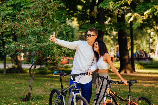Young stylish couple in casual outfits are taking selfie on a smartphone in the city park, holding their bicycles. Cyclists are taking photos on the phone.