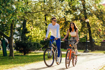 Young romantic couple of cyclists is biking in the summer park, smiling and heeling happy. Attractive caucasian man is looking at his girlfriend with pleasure.