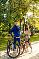 Vertical photo of stylish smiling positive couple standing in the park with sport bikes. Sunshine and greenery on the background.