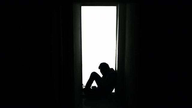 Silhouette shot: a sad, desperate man, sitting on the floor near a door in his house, alone and crying, letting his bad feelings come out.
