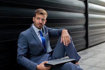 Portrait of handsome businessman in suite sitting at the floor in the front of the modern building. 