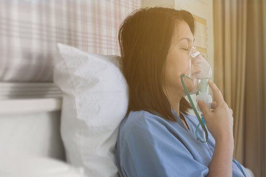 Sick beautiful female in blue cloth hold nasal mask with respiratory problem in hospital room. Asian woman patient inhalation therapy by the mask of inhaler with soft stream smoke from bronchodilator.