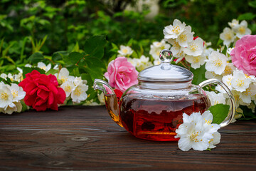 Obraz na płótnie Canvas Glass teapot among flowering bushes of roses and jasmine. Outdoor, picnic, brunch. Green leaves background in blur