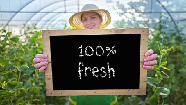 A woman farmer invites buying organic vegetables and fruits. Agriculture. Smiling woman with a sign in her hands calls for healthy food. Vegetarianism, vegan. Greenhouse, family vegetable growing busi