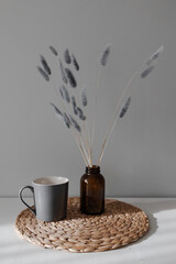 still life with grey cup and dry grey flowers in the brown vase on on the rattan napkin near grey wall. 