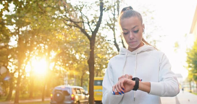 Attractive Caucasian woman in earphones listening to music and setting up stop watch. Adult female athlete timing jogging and trying to beat record in street. Sport, workout, training concept.