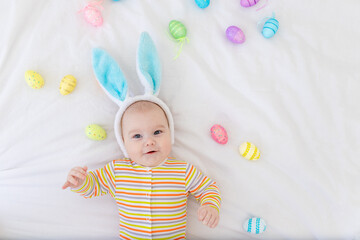 happy baby boy with rabbit ears on his head lying on the bed with Easter eggs, cute funny smiling little baby. The concept of Easter. Space for text
