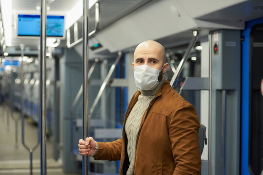 A man with a beard in a medical face mask to avoid the spread of COVID-19 is standing and holding the handrail in a subway car. A bald guy in a surgical mask is keeping social distance on a train.
