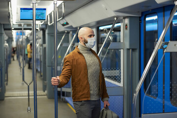 A man with a beard in a medical face mask to avoid the spread of coronavirus is riding a modern subway car. A bald guy in a surgical mask is keeping social distance on a train.