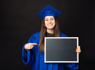 Cheerful young student woman wearing bachelor and pointing at black chalk board