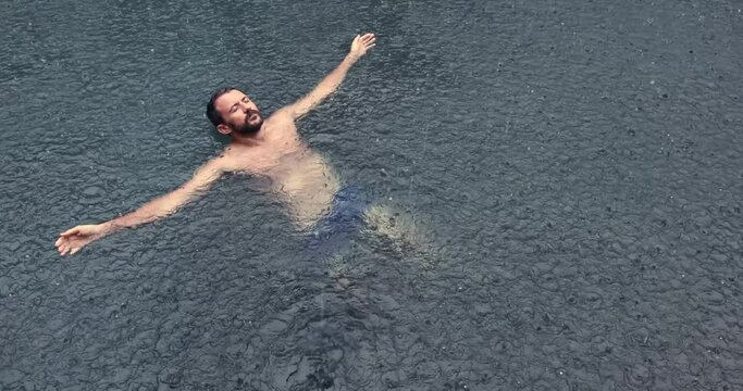 Young man floating on the back in the dark water of simming pool, ocean or lake under the rain drops