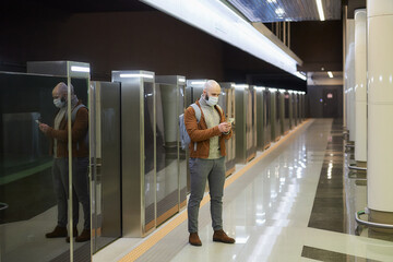 A full-length photo of a man in a face mask who is scrolling the smartphone while waiting for a train at the subway platform. A bald guy in a surgical mask is keeping social distance.